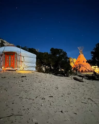 Illuminated yurt and teepee on sandy ground at night with starry sky and rocky cliff backdrop