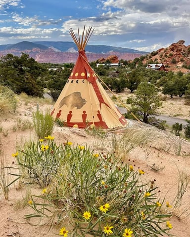 Decorated teepee in desert landscape with red rock formations, scattered houses, and yellow wildflowers in foreground