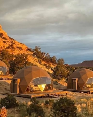 Four geodesic dome glamping structures nestled against red rock cliffs at sunset in desert landscape.