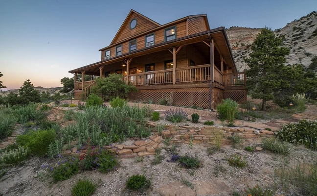 Wooden farmhouse with wraparound porch and desert landscaping at dusk, rocky hillside backdrop