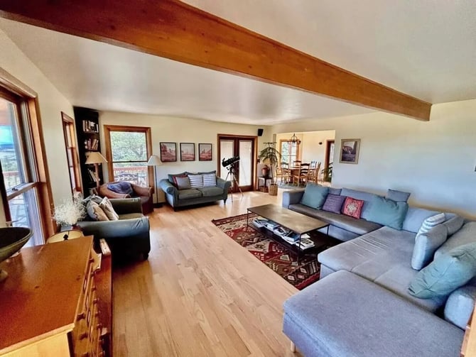Spacious open-plan living room with exposed wooden beam, multiple seating areas, hardwood floors, and views to dining area and deck.