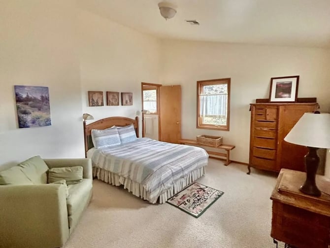 Spacious bedroom with wooden bed, striped bedding, green armchair, wooden dresser, nightstands, and windows with natural light