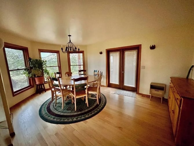 Bright dining room with wood table and chairs on round rug, multiple windows with wood frames, chandelier, and hardwood floors.