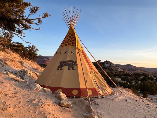 Decorated teepee on sandy desert hillside with red rock formations and sparse vegetation in background