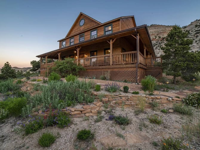 Wooden farmhouse with wraparound porch and elevated foundation in desert landscape with native plants and rocky hillside.